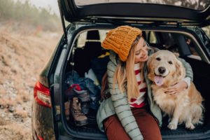 Photo of a young smiling woman and her dog sitting the trunk of a car on a beautiful autumn day; taking a short break during their road trip.